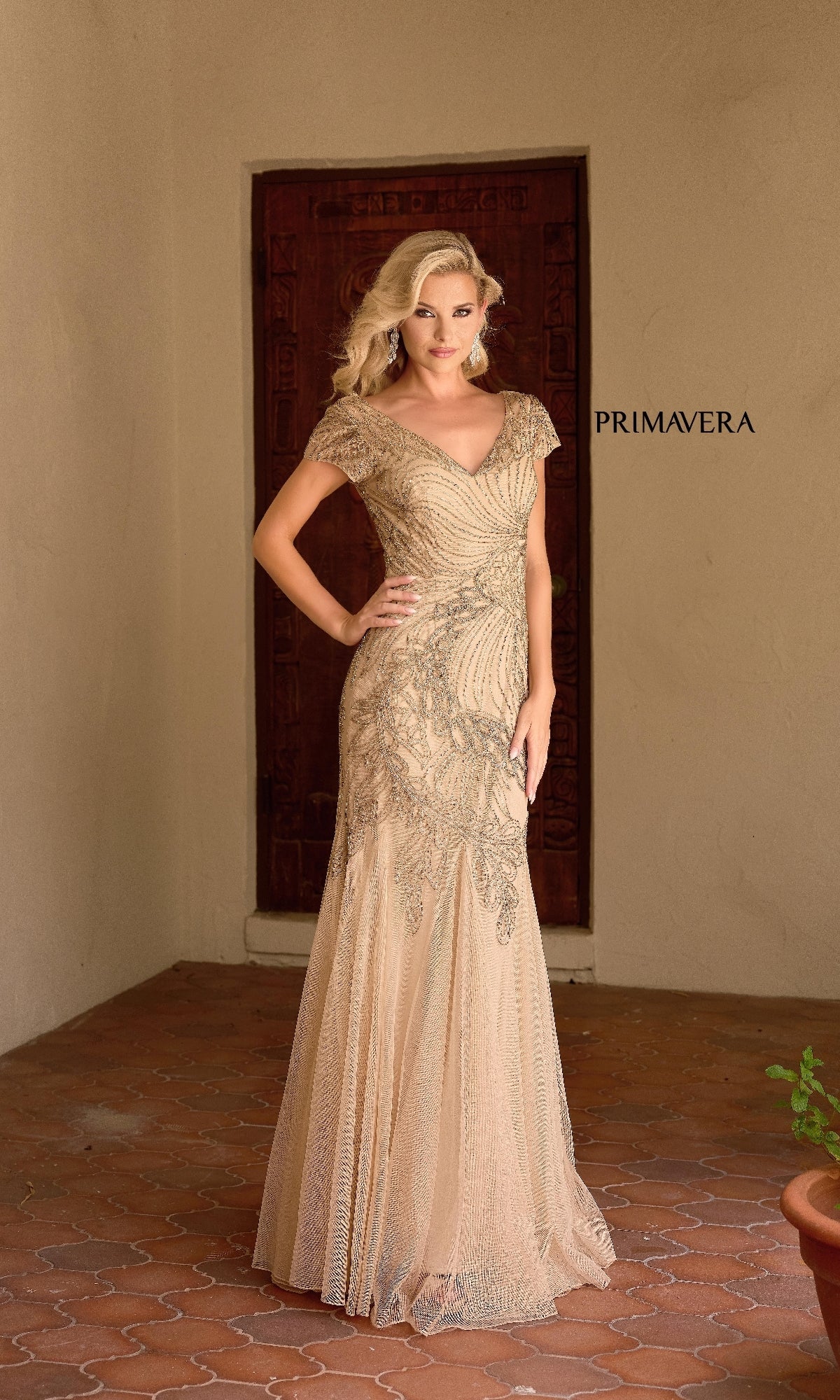 A woman stands on a tiled patio in the Primavera 13148 long formal dress, featuring beading embellishments and cap sleeves.